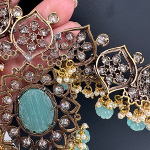 May include: Close-up of an ornate necklace with a gold and silver finish. The necklace features a large, oval, light blue stone surrounded by clear crystals. Small white pearls and light blue beads dangle from the necklace.