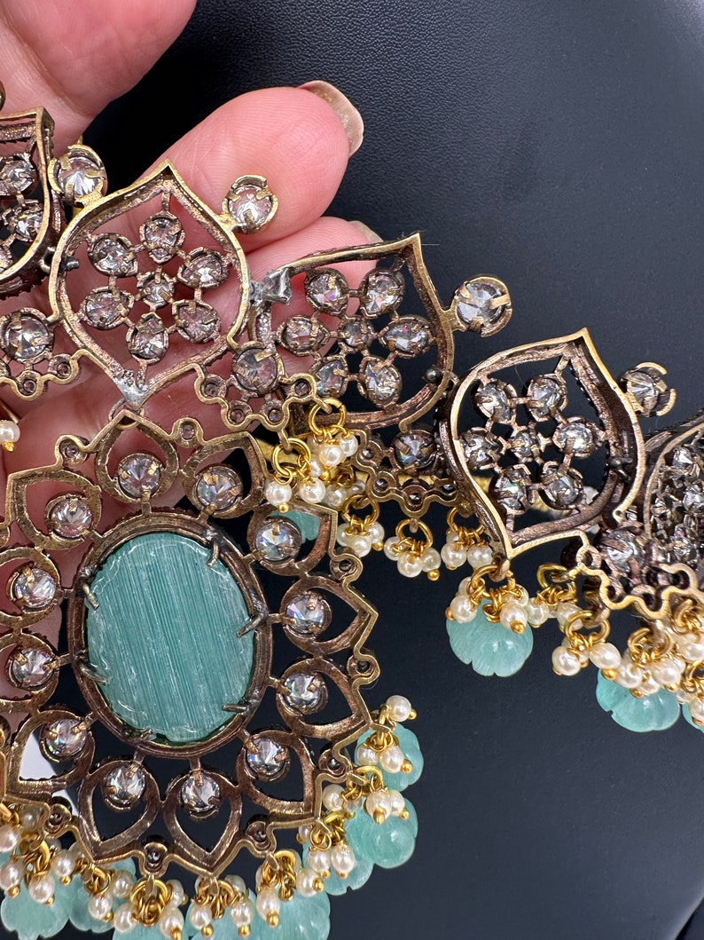May include: Close-up of an ornate necklace with a gold and silver finish. The necklace features a large, oval, light blue stone surrounded by clear crystals. Small white pearls and light blue beads dangle from the necklace.