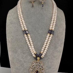 May include: A pearl necklace and earring set displayed on a mannequin. The necklace features two strands of white pearls, gold accents, and a decorative pendant. The earrings and pendant have a floral design with clear and purple gemstones.
