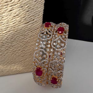 May include: Two ornate gold-toned bangles with intricate floral and leaf patterns. The bangles are encrusted with clear, sparkling crystals and feature large, oval-shaped, ruby-red gemstones. The jewelry is displayed on a white surface.