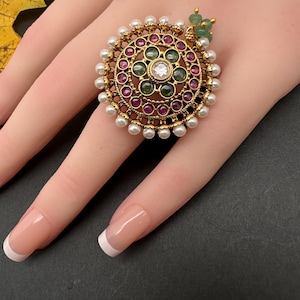 May include: A gold-toned ring with a circular design, featuring a central clear stone surrounded by green and red gemstones. The ring is further embellished with small white pearls around the outer edge. A cluster of green beads accents the side.
