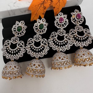May include: Four pairs of ornate, silver-toned earrings with clear and colored gemstones. Each pair features a floral top, a curved, openwork design, and a bell-shaped bottom. Two pairs have green gemstones, and two have red gemstones.
