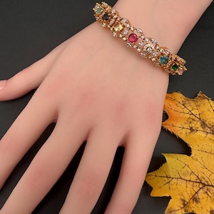 May include: Gold-toned bracelet with an intricate design, featuring a series of colorful gemstones including red, blue, green, and yellow. The bracelet is set with smaller, clear stones, creating a sparkling effect. The bracelet is displayed on a hand.