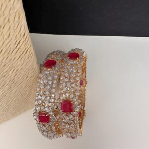 Peut inclure: Deux bracelets dorés incrustés de cristaux clairs et étincelants et de pierres précieuses rouges carrées vibrantes. Les bijoux sont exposés sur une surface blanche avec un fond neutre, mettant en valeur les détails complexes des bracelets.