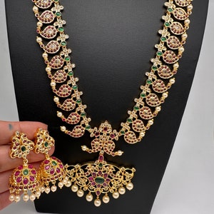 May include: A gold-toned jewelry set featuring a long necklace and matching earrings. The necklace and earrings are adorned with red, green, and clear gemstones, along with small white pearls. The necklace has a central pendant with a floral design.