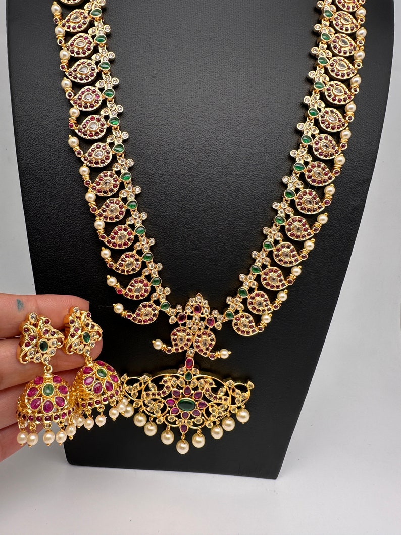 May include: A gold-toned jewelry set featuring a long necklace and matching earrings. The necklace and earrings are adorned with red, green, and clear gemstones, along with small white pearls. The necklace has a central pendant with a floral design.