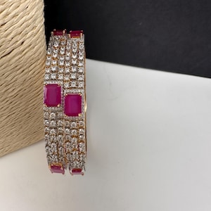 May include: Two gold-toned bangles with a repeating pattern of small, clear stones and larger, rectangular, magenta-colored stones. The bangles are displayed on a white surface with a black background.