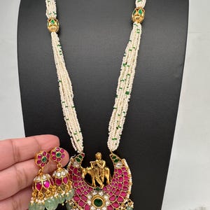 May include: An ornate gold-toned necklace and earring set. The necklace features a pendant with a figure playing a flute, surrounded by pink and green gemstones. The earrings have a floral design with pink and green accents, and dangling green beads.
