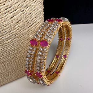 May include: Gold-toned bangles with alternating clear and ruby-colored stones. The bangles are stacked, showcasing the intricate design and the contrast between the gold metal and the vibrant gemstones. The background is neutral, highlighting the jewelry.