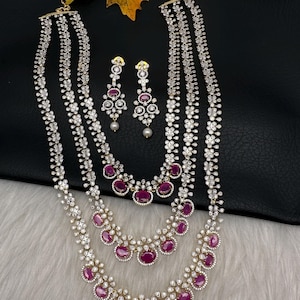 May include: A three-strand necklace and matching earrings set. The necklace features clear, round stones and ruby-colored oval stones. The earrings have a similar design with a dangling pearl. The jewelry is set against a black background.