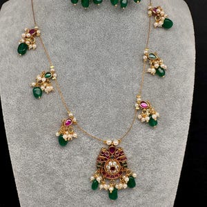 May include: A gold-toned jewelry set featuring a necklace and matching earrings. The set includes ruby-colored and clear stone accents, with dangling green beads and small white pearls. The necklace has a central pendant with a similar design.