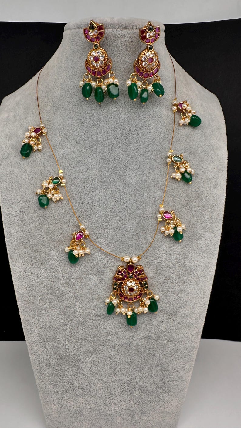 May include: A gold-toned jewelry set featuring a necklace and matching earrings. The set includes ruby-colored and clear stone accents, with dangling green beads and small white pearls. The necklace has a central pendant with a similar design.