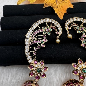 May include: Gold-toned earrings with intricate floral and leaf designs. The earrings feature clear and ruby-colored gemstones, with a pearl drop. The design includes a floral ear cuff and a dangling, gem-studded teardrop shape.