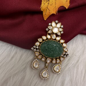 May include: A gold-toned pendant featuring a central oval-shaped green stone with an intricate floral carving. The pendant is surrounded by small, clear, faceted stones arranged in a flower-like pattern at the top and three dangling teardrop-shaped stones at the bottom.
