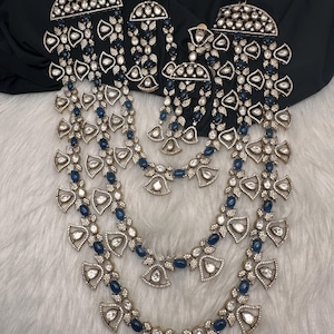 May include: An ornate jewelry set featuring a necklace, earrings, and a headpiece. The pieces are silver-toned with clear and dark blue gemstones. The necklace has multiple strands, and the earrings and headpiece have a matching design. The set is displayed on a white, fluffy surface.
