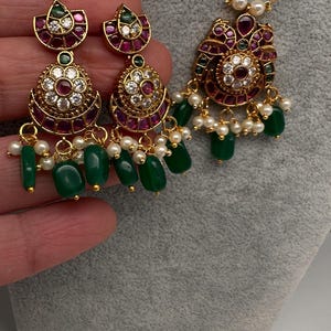 May include: A gold-toned jewelry set featuring a pendant necklace and matching earrings. The pieces are adorned with red and clear gemstones, white pearls, and green beads. The earrings and pendant have a floral design.