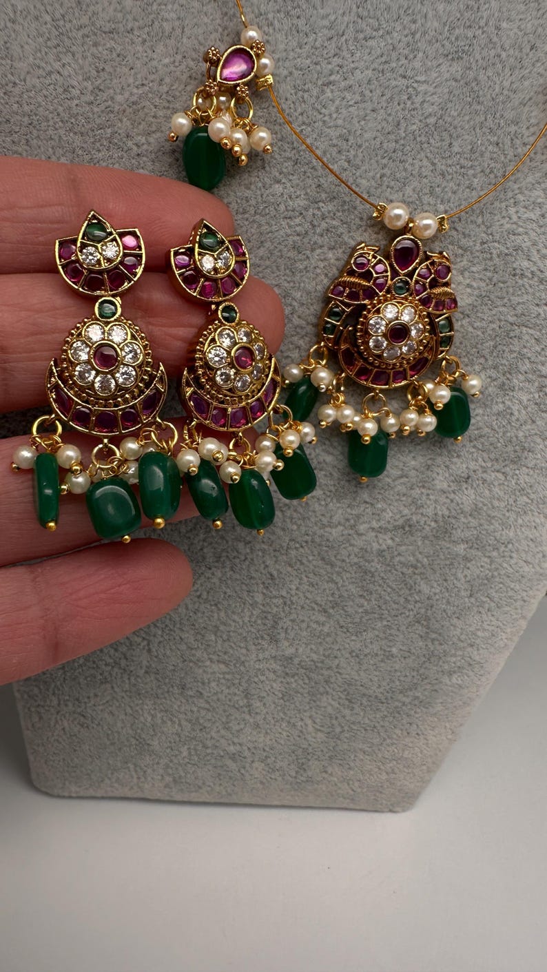 May include: A gold-toned jewelry set featuring a pendant necklace and matching earrings. The pieces are adorned with red and clear gemstones, white pearls, and green beads. The earrings and pendant have a floral design.