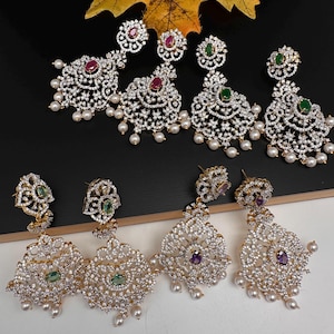 May include: An assortment of ornate, dangling earrings with a gold-tone setting. The earrings feature clear, sparkling stones, and colored gemstones in red, green, and purple. Each earring has a cluster of small, white pearls at the bottom.