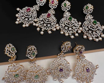 Puede incluir: Una variedad de pendientes colgantes ornamentados con un engaste dorado. Los pendientes presentan piedras claras y brillantes, y piedras preciosas de colores en rojo, verde y morado. Cada pendiente tiene un grupo de pequeñas perlas blancas.