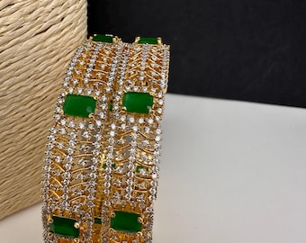 Puede incluir: Dos brazaletes dorados con un patrón repetido de cristales transparentes y piedras rectangulares verde esmeralda. Los brazaletes se exhiben sobre una superficie blanca con un fondo beige texturizado.