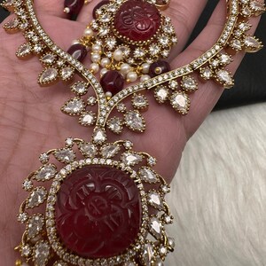 May include: An ornate gold-coloured jewellery set, comprising a necklace and earrings. The set features large, carved red gemstones, surrounded by clear, teardrop-shaped crystals. Small pearls and red beads add to the elegant design.