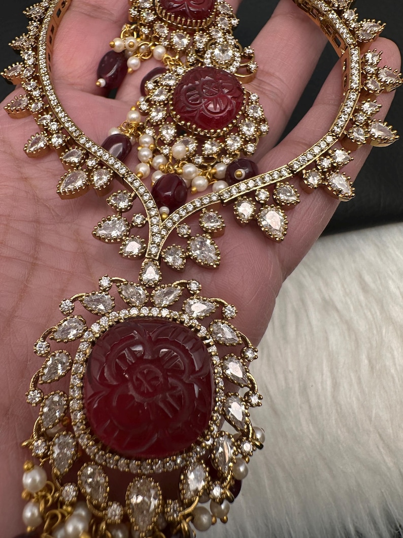 May include: An ornate gold-coloured jewellery set, comprising a necklace and earrings. The set features large, carved red gemstones, surrounded by clear, teardrop-shaped crystals. Small pearls and red beads add to the elegant design.