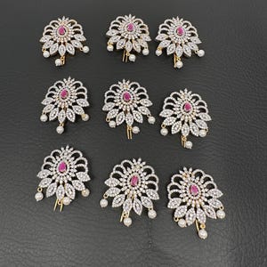 May include: Nine ornate hair clips with a floral design. Each clip features a central red gemstone surrounded by clear crystals and dangling faux pearls. The clips are arranged on a dark surface.
