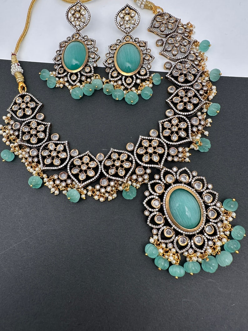 May include: An ornate jewelry set featuring a necklace and matching earrings. The set includes a necklace with a large oval pendant and a pair of earrings, each with a large oval stone. The jewelry is embellished with small white stones and light green beads.
