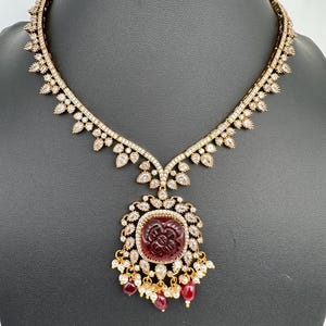 May include: A gold necklace with a large, square, burgundy pendant. The pendant has a carved floral design and is surrounded by clear gemstones. The necklace has a delicate, ornate design with clear gemstones and small burgundy beads.