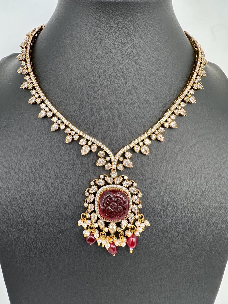May include: A gold necklace with a large, square, burgundy pendant. The pendant has a carved floral design and is surrounded by clear gemstones. The necklace has a delicate, ornate design with clear gemstones and small burgundy beads.