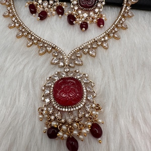 May include: A gold-coloured necklace and earring set. The necklace features a large, round, red gemstone pendant surrounded by clear crystals. The earrings and necklace have a floral design with dangling red beads and small white pearls.
