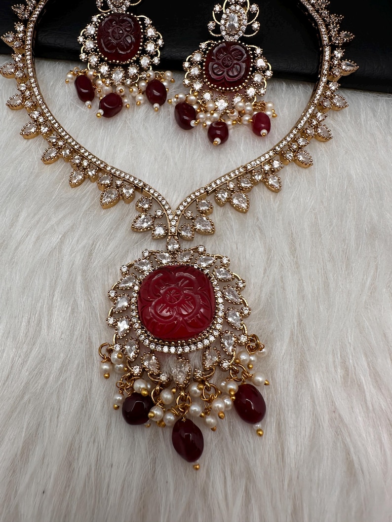 May include: A gold-coloured necklace and earring set. The necklace features a large, round, red gemstone pendant surrounded by clear crystals. The earrings and necklace have a floral design with dangling red beads and small white pearls.