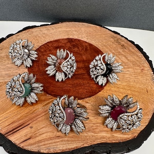 May include: Six flower-shaped rings with various center stones, including clear, black, green, and pink. The rings are set on a wooden surface. The rings have a silver-colored metal setting and are encrusted with small, clear stones.