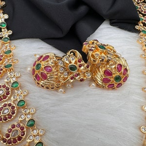 May include: Gold-toned jewelry set featuring earrings and a necklace. The earrings and necklace are adorned with red, green, and clear gemstones. The jewelry is displayed on a white, fluffy surface with a black fabric backdrop.