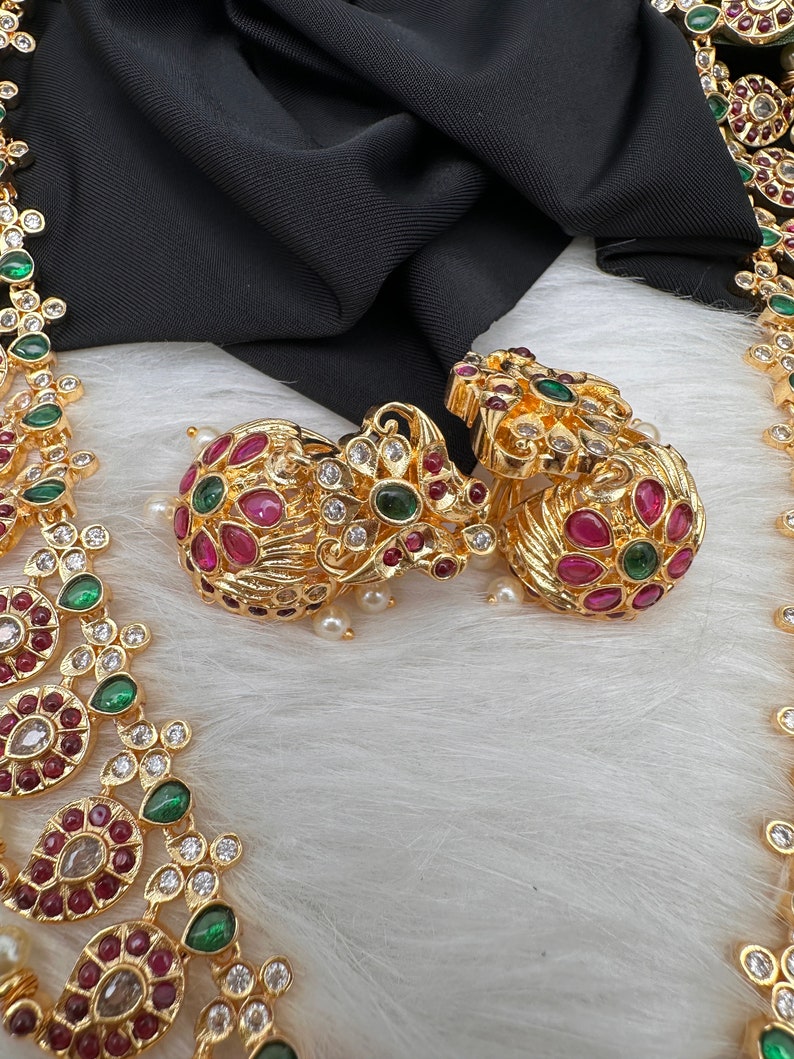 May include: Gold-toned jewelry set featuring earrings and a necklace. The earrings and necklace are adorned with red, green, and clear gemstones. The jewelry is displayed on a white, fluffy surface with a black fabric backdrop.