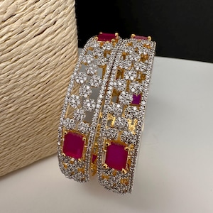 May include: Two gold-coloured bangles with intricate floral and geometric patterns. The bangles are encrusted with clear, sparkling stones and feature square, ruby-red accents. The jewellery is displayed on a white surface.