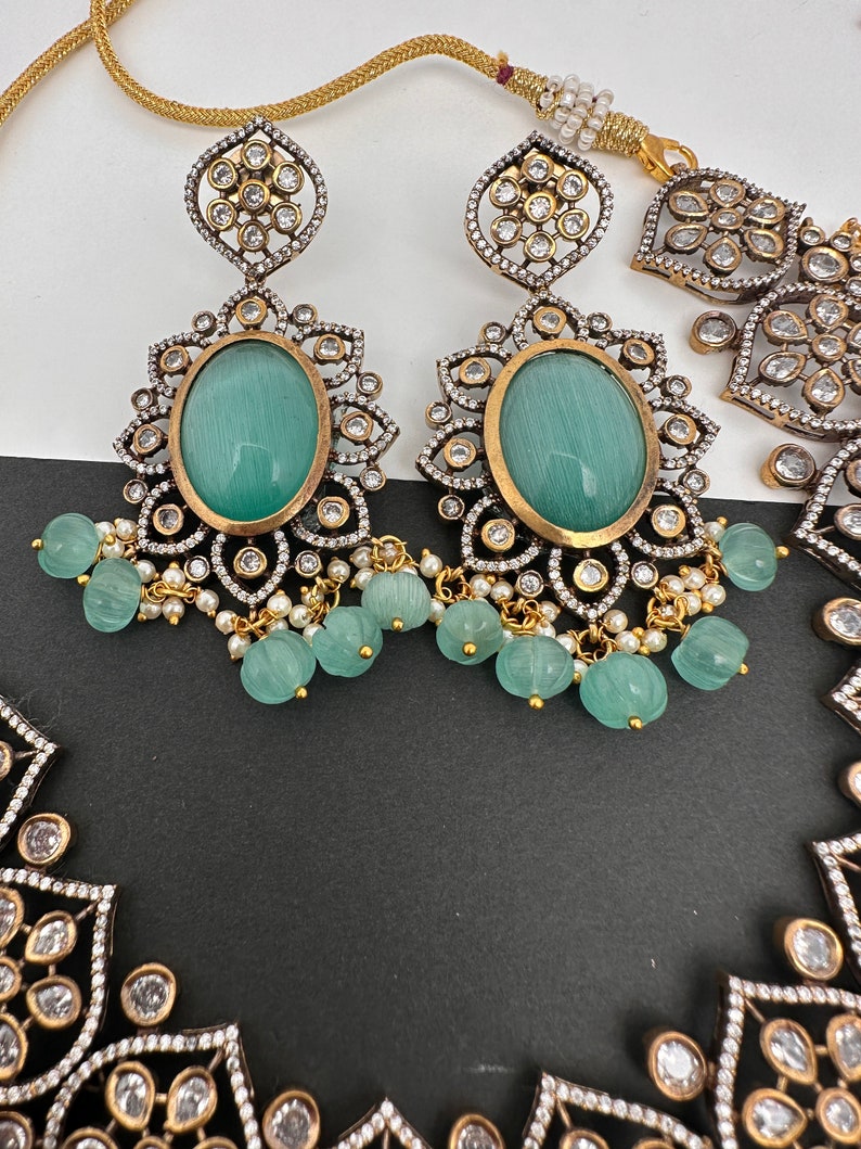 May include: Close-up of an ornate jewelry set featuring earrings and a necklace. The earrings have large oval, light green stones, surrounded by gold and silver filigree, and dangling green beads. The necklace has a similar design.