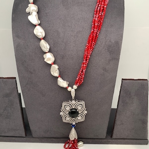 May include: A statement necklace featuring large, irregular white pearls strung on red cord, paired with strands of small red beads. The pendant is a silver-toned, floral-shaped design with a black center stone and dangling white pearls.