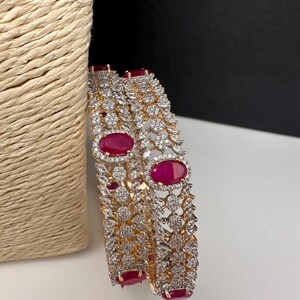 May include: Two gold bangles with detailed patterns. Each bangle has a large, oval ruby-red stone at the centre, encircled by smaller, clear, sparkling stones. The design incorporates floral and leaf-like motifs.