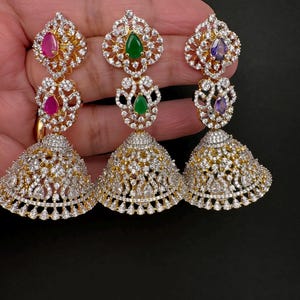 May include: Three gold-coloured, ornate earrings with a bell shape. Each earring features a cluster of clear crystals and a coloured gemstone: pink, green, and purple. The earrings are designed with intricate details and a sparkling finish.