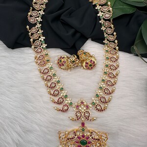 May include: A gold-toned necklace and matching earrings set. The necklace features a long chain with paisley-shaped pendants adorned with red and green gemstones, and white pearls. The earrings are round with similar red and green stones.