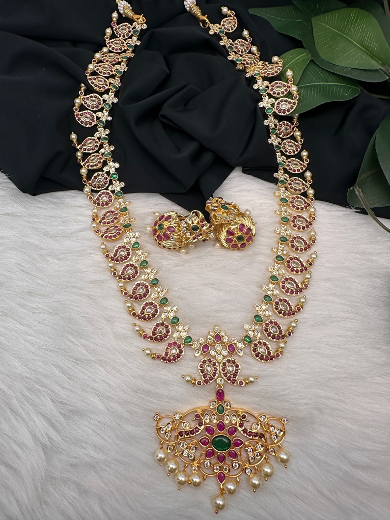 May include: A gold-toned necklace and matching earrings set. The necklace features a long chain with paisley-shaped pendants adorned with red and green gemstones, and white pearls. The earrings are round with similar red and green stones.