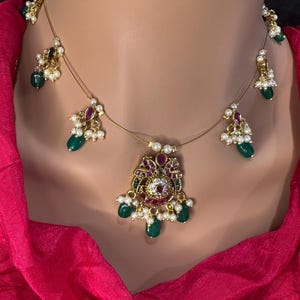 May include: A gold-toned necklace with a central pendant and dangling accents. The pendant features a floral design with red and white gemstones, surrounded by green teardrop-shaped stones and small pearls. The necklace is displayed on a mannequin.