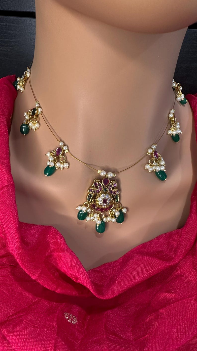 May include: A gold-toned necklace with a central pendant and dangling accents. The pendant features a floral design with red and white gemstones, surrounded by green teardrop-shaped stones and small pearls. The necklace is displayed on a mannequin.