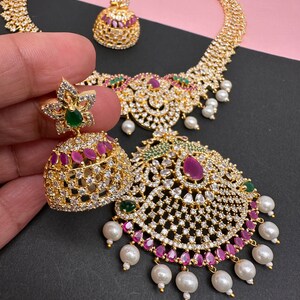 Op de afbeelding: Een goudkleurige sieradenset met een ketting en oorbellen. De ketting en oorbellen zijn versierd met heldere en robijnkleurige stenen en witte parels. De oorbellen hebben een bloemenontwerp met een groene steen in het midden.
