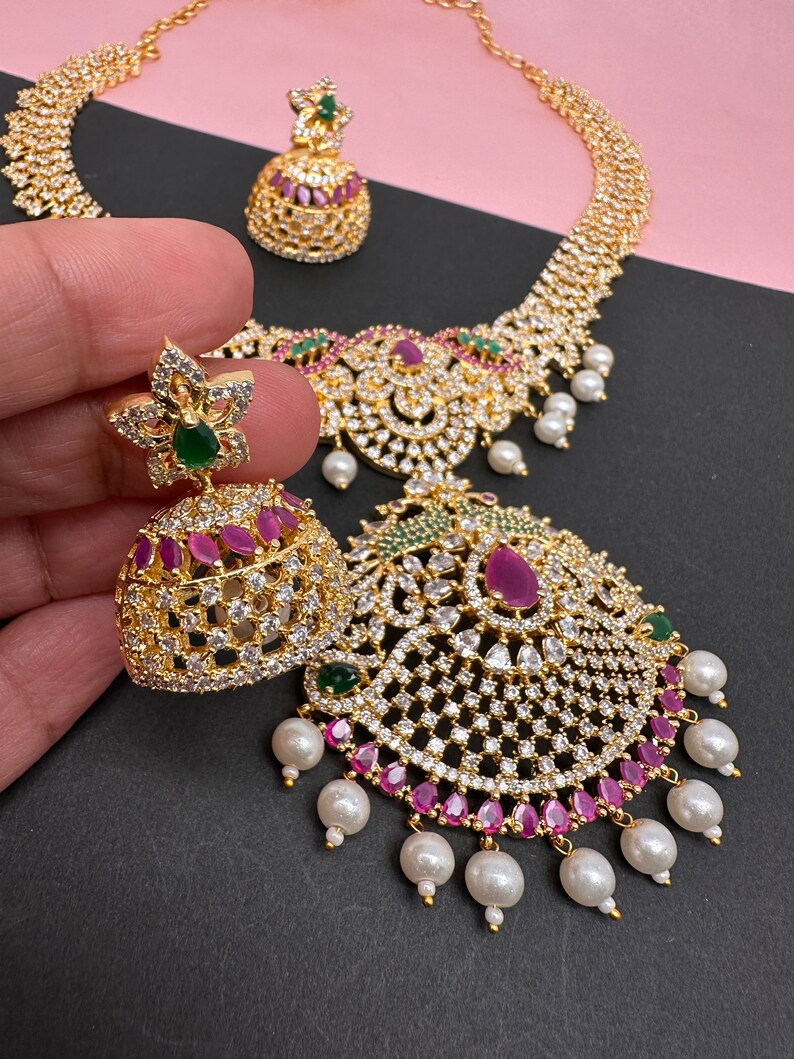 Op de afbeelding: Een goudkleurige sieradenset met een ketting en oorbellen. De ketting en oorbellen zijn versierd met heldere en robijnkleurige stenen en witte parels. De oorbellen hebben een bloemenontwerp met een groene steen in het midden.