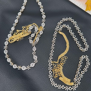 May include: Two necklaces with a gold chain and white gemstones. The necklaces are arranged on a black surface. One necklace has a teardrop design, and the other has a floral design. The necklaces are elegant and suitable for special occasions.