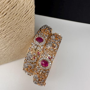 May include: Two gold-toned bangles with intricate filigree and sparkling clear stones. Each bangle features a large, oval-shaped ruby-red stone at the center. The design includes smaller red stones and detailed patterns.