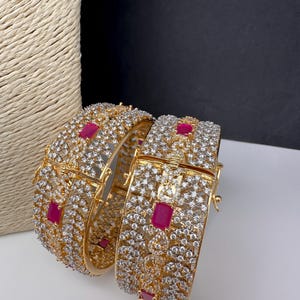 May include: Two gold-toned bangles encrusted with clear crystals and accented with rectangular ruby-red stones. The bangles have an openwork design and a hinged clasp. The background is a neutral white and black.