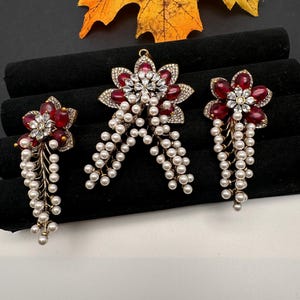 May include: A jewelry set featuring a flower design with red gemstone petals, clear crystal accents, and dangling faux pearls. The set includes a pendant, earrings, and a ring, all with a gold-tone metal finish. The background is black and white.